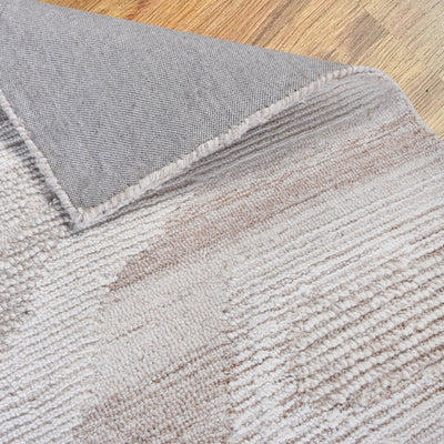 Close-up of the corner of an ivory and beige hand-tufted rug, made of 100% wool, folded to show the backing and layered pile texture on a wood floor.