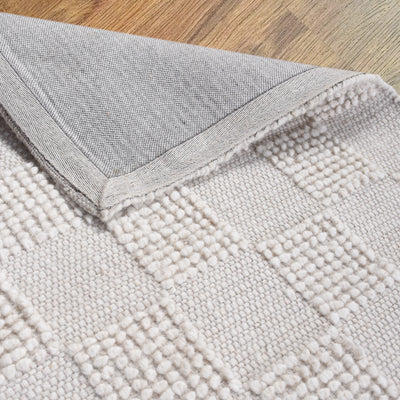 Ivory hand-woven wool rug, featuring a geometric pattern with the corner folded back to show the underside, on a floor background.