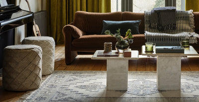 Warm living room with a brown corduroy sofa, patterned throw, and blue cushion, paired with two cube pouffes, a soft patterned rug, and two stone pedestal coffee tables styled with candles, books, and a glass vase of flowers.