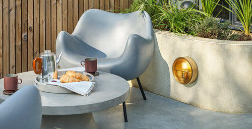 Outdoor decorative wall light with a brass finish, styled in a modern patio seating area with concrete table, chairs, and greenery.