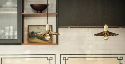 Pair of brass pendant lights with pleated shades and exposed bulbs hanging above a modern rustic kitchen with white tiled walls and dark cabinets.