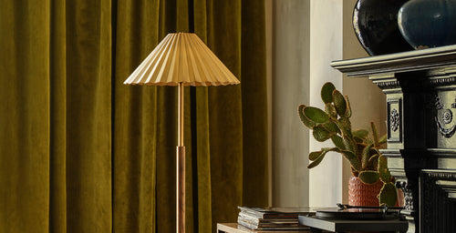 Modern floor lamp with a pleated shade and slim brass and wood base, styled beside a black fireplace and green velvet curtains.