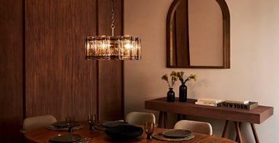 Modern dining room with a statement glass chandelier casting a warm glow above a wooden table, styled with neutral chairs, black vases, and contemporary decor.