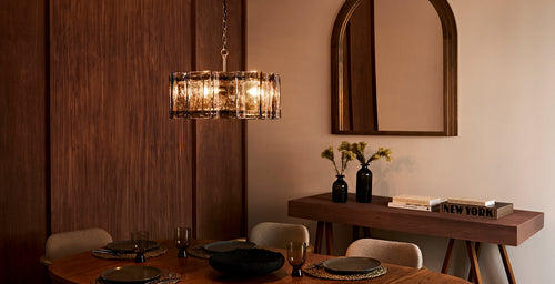 Modern dining room with a statement glass chandelier casting a warm glow above a wooden table, styled with neutral chairs, black vases, and contemporary decor.