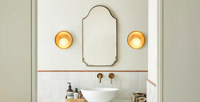 Modern bathroom with twin brass wall lights flanking a vintage mirror above a wooden vanity unit with a white countertop basin.
