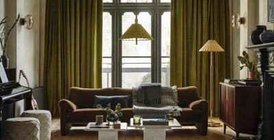 A warm living room with olive-green curtains, a brown sofa, marble coffee tables, and pleated lighting creating a cosy, vintage feel.