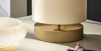 Modern table lamp with a textured brass base and cream fabric shade, styled on a neutral stone surface.