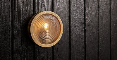 Outdoor commercial bulkhead wall light in solid brass with ribbed glass, mounted on dark timber cladding with water droplets.