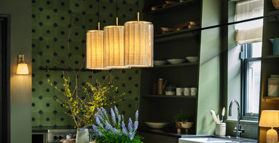 Modern kitchen with layered lighting, featuring a trio of cylindrical pendant lights above the island, a wall sconce with a warm shade, and a small table lamp beside the sink.