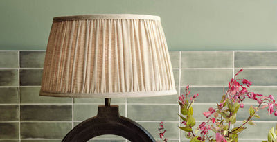 Pleated fabric lampshade in a soft neutral tone, styled on a decorative table lamp with green tiled wall backdrop and floral accents.