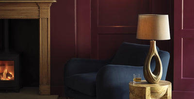 Ornate wooden ellipse table lamp with fabric shade on a marble side table, styled beside a navy armchair and fireplace.