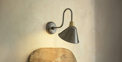 Swan neck wall light with pewter cone shade and brass detailing, mounted on a neutral wall above rustic wooden decor.