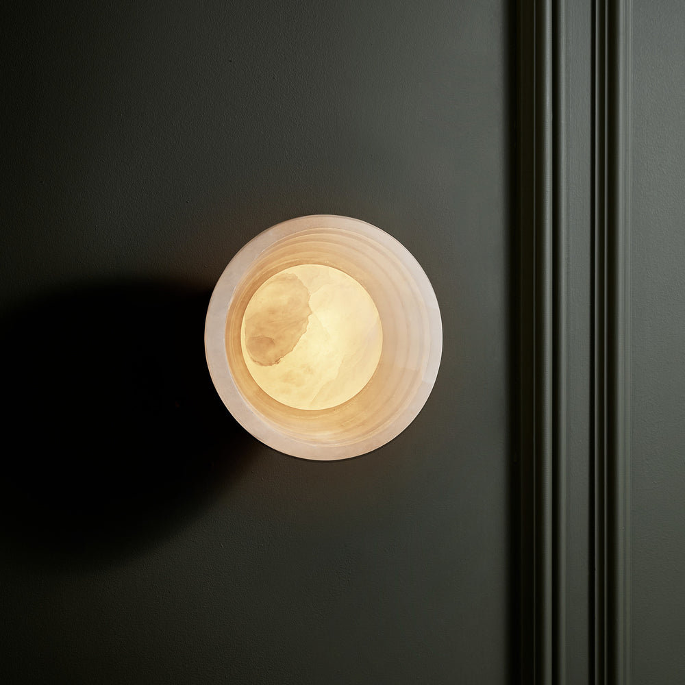 Corby Alabaster Stepped Diffuser Wall Light  6 Inch with Aged Brass Holder lit up on a dark wall
