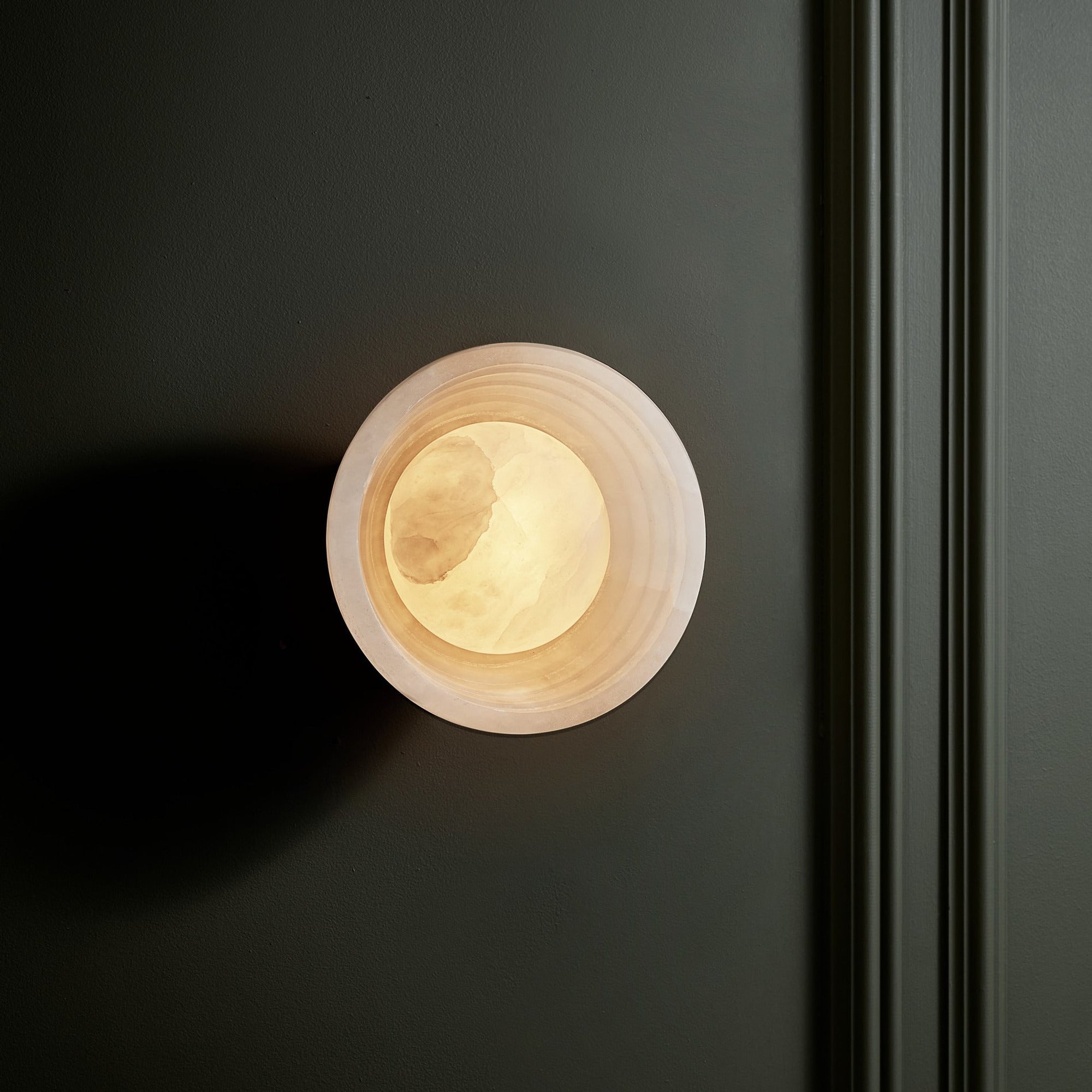 Corby Alabaster Stepped Diffuser Wall Light  6 Inch with Aged Brass Holder lit up on a dark wall