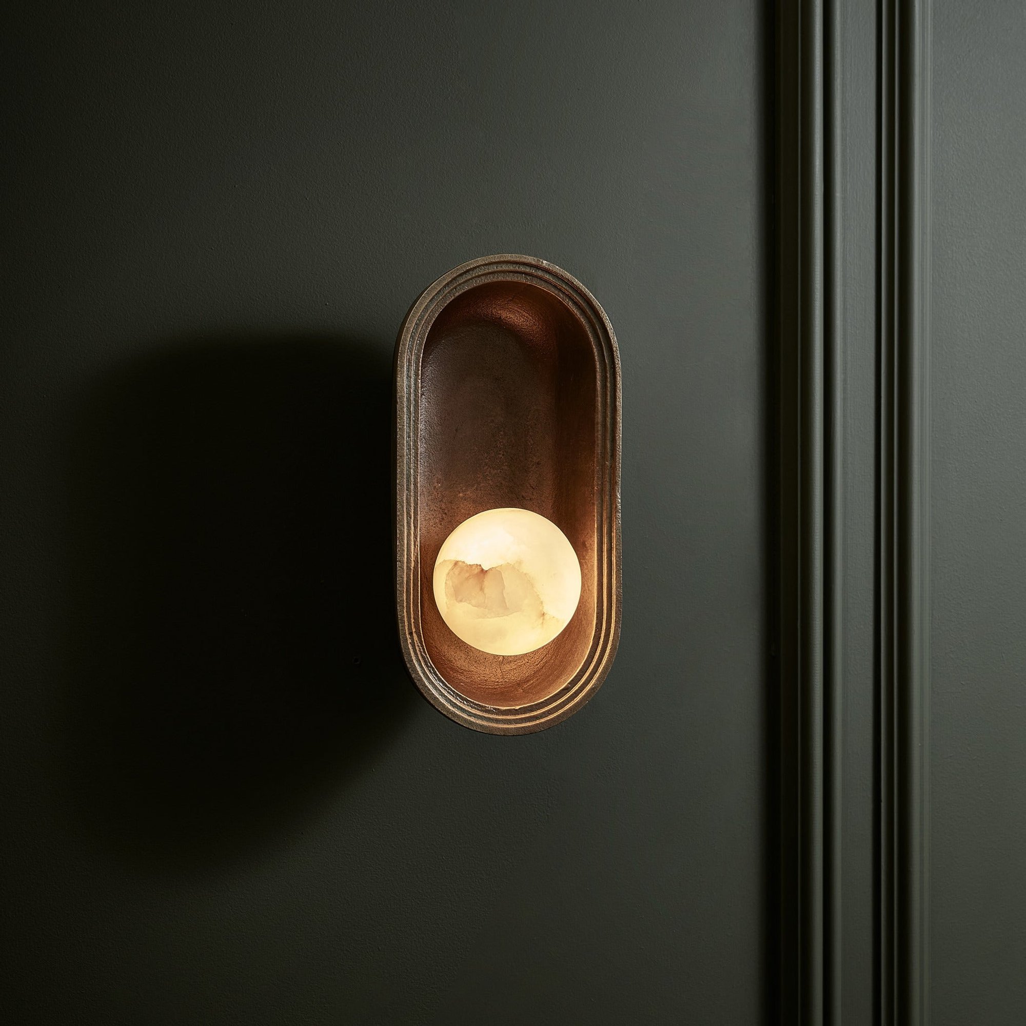 Corby Cast & Alabaster Oval Wall Light 5 Inch in Aged Brass Lit Up on a dark wall